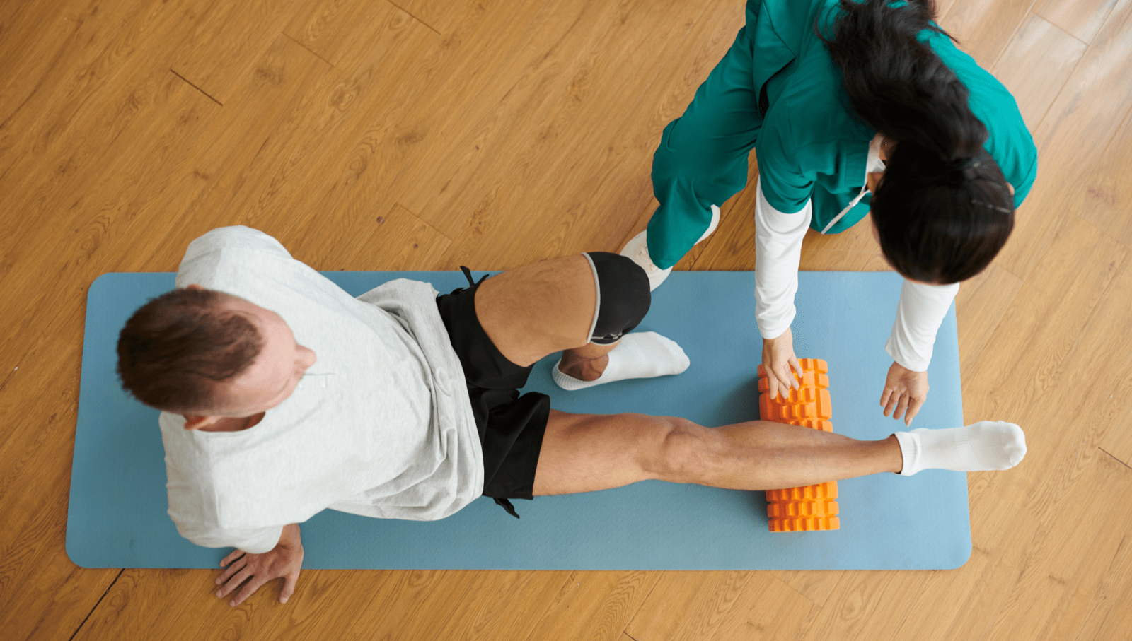 A physiotherapist teaching a patient how to use a foam roller on their calf to relieve tension while sitting on a yoga mat